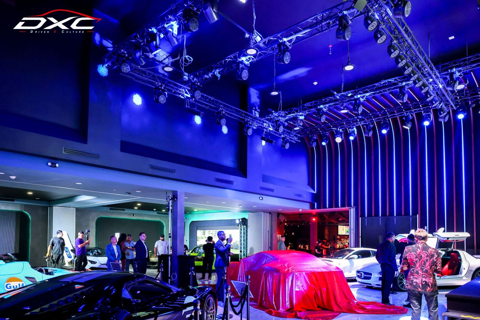The DXC Launch Night showroom under blue stage lights, with show cars on the floor and one under a red cover