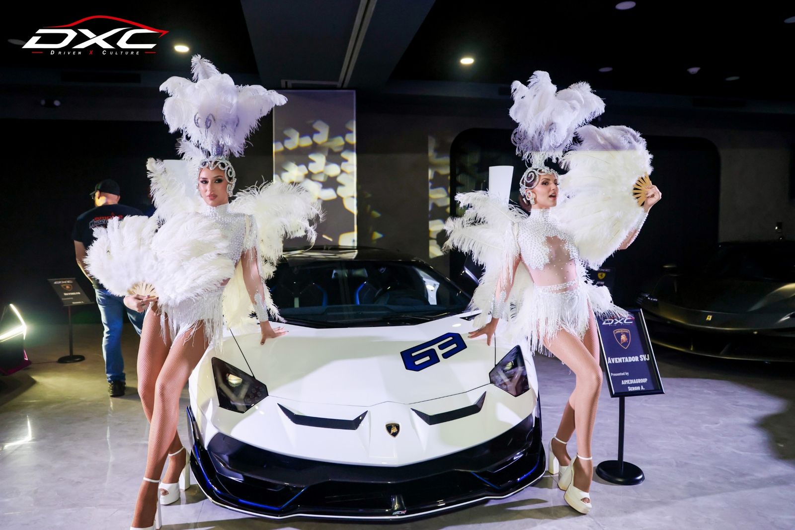 Two showgirls in white feathers posing with a white Lamborghini Aventador SVJ at DXC Launch Night